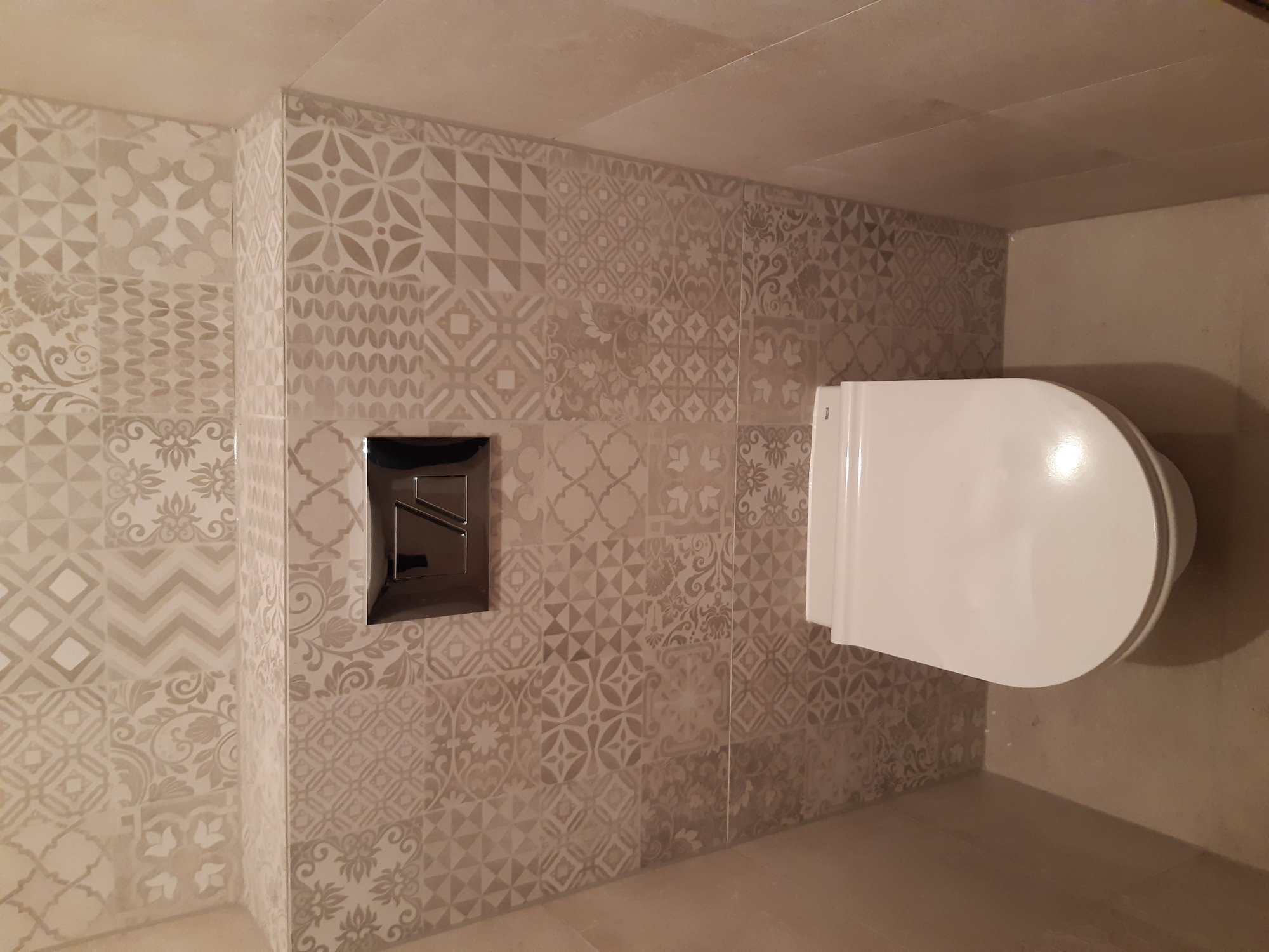 Wykończona łazienka z toaletą i mozaiką z płytek w odcieniach beżu i szarości, chromowany panel spłukujący.