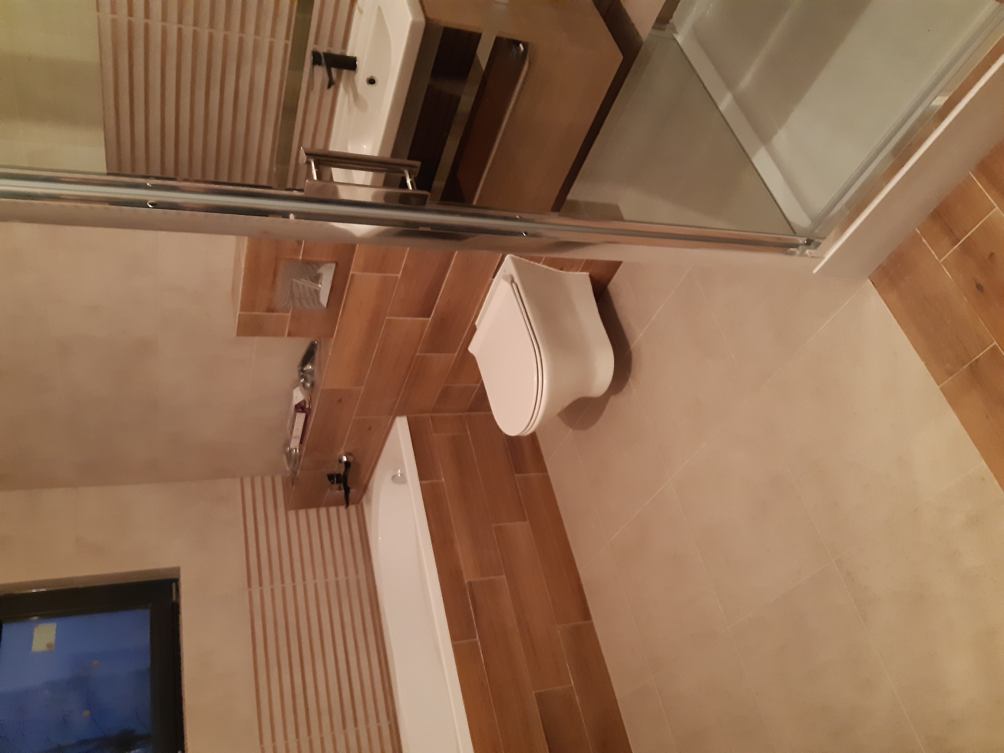 Nowoczesna łazienka z drewnopodobnymi płytkami na ścianach i podłodze, białą armaturą, kabiną prysznicową ze szklanymi drzwiami, umywalką z szafką i toaletą.