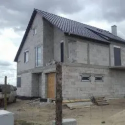 Surowy, dwukondygnacyjny budynek z betonu komórkowego w Kłodawie, z ciemnym dachem i widocznymi otworami okiennymi oraz drzwiowymi, w trakcie budowy, na tle pochmurnego nieba.