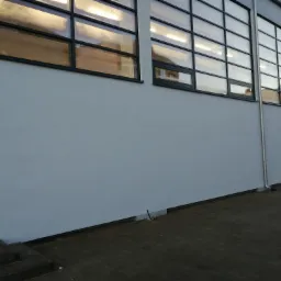 Świeżo otynkowana szara ściana budynku z rzędem okien z czarnymi ramami i widoczną instalacją odprowadzającą wodę deszczową.