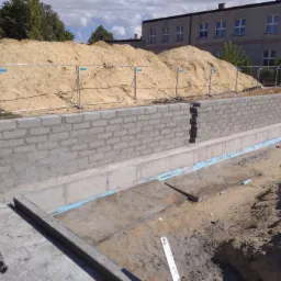 Wznoszenie muru z bloczków betonowych na fundamencie, widoczne zabezpieczenia wykopu, w tle budynek i hałdy piasku.