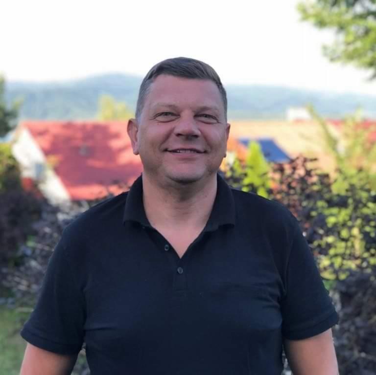 Uśmiechnięty mężczyzna w czarnej koszulce polo na tle domu z czerwoną dachówką i zieleni, portret w plenerze, naturalne światło.