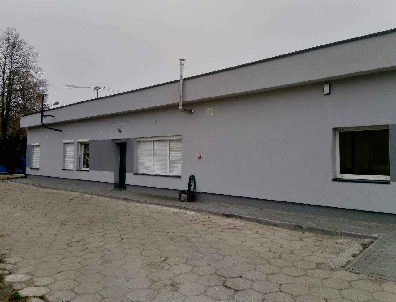 Szary budynek z oknami z żaluzjami, rurą kominową i wężem ogrodowym zwiniętym w koszyku przy wejściu, widoczny z zewnątrz na tle brukowanej powierzchni.