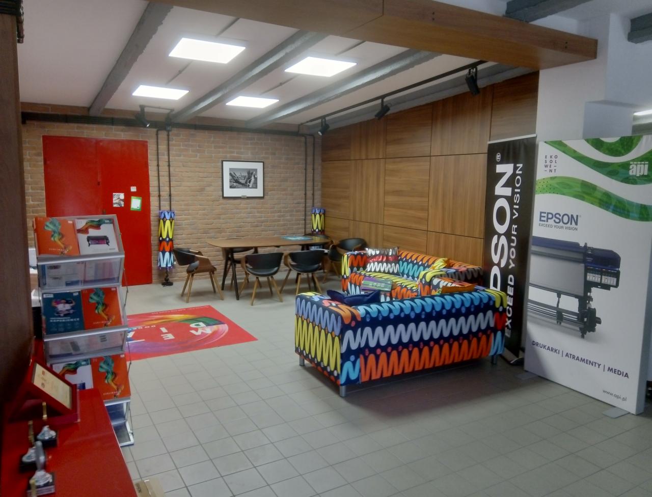 Wnętrze biura z kolorowymi sofami i banerami reklamowymi drukarki Epson, widoczna ceglana ściana i drewniane panele ścienne.