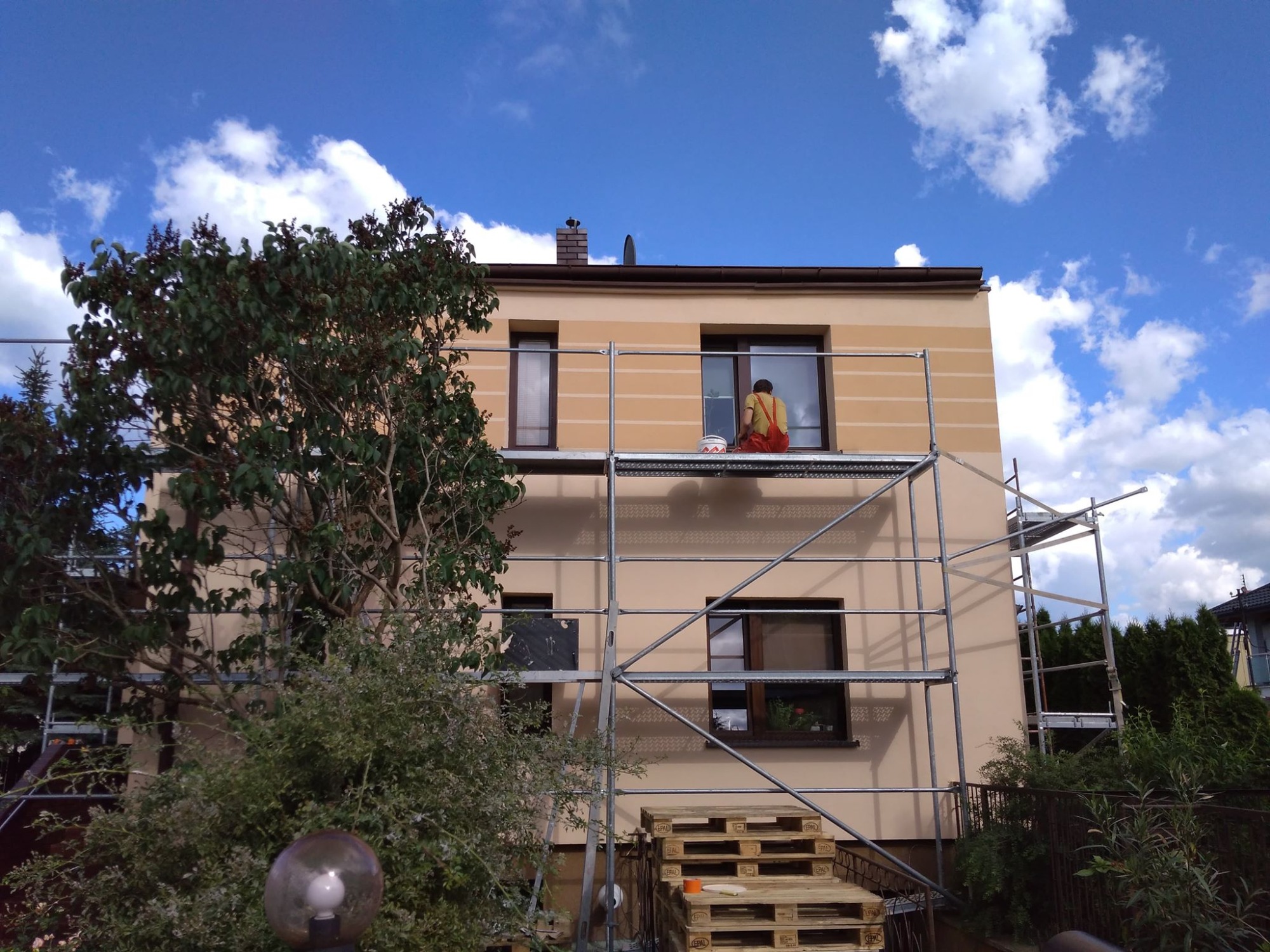 Dwupiętrowy dom jednorodzinny z beżową elewacją w trakcie prac, widoczny pracownik w czerwonym kombinezonie na rusztowaniu, malujący pasy na ścianie, obok puszka z farbą, w tle błękitne niebo...