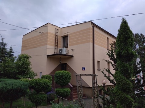 Dwukondygnacyjny budynek z jasną elewacją w odcieniach beżu, z poziomymi pasami w jaśniejszym odcieniu; drabina oparta o ścianę przy balkonie, widoczny klimatyzator; zadbany ogród z krzewami...