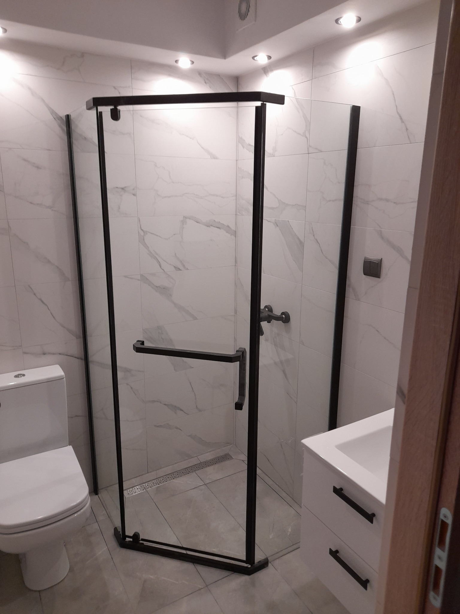 Nowoczesna kabina prysznicowa z czarnymi profilami, szklanymi ściankami i odpływem liniowym w podłodze, na tle płytek imitujących marmur, widoczna również umywalka z szafką i toaleta.