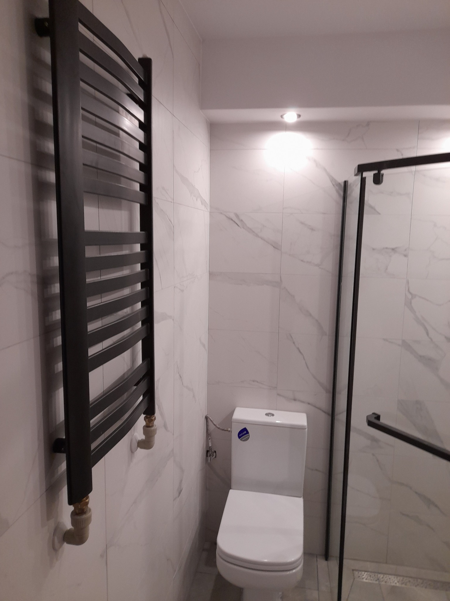 Nowoczesna łazienka z marmurowymi płytkami, czarnym grzejnikiem drabinkowym, toaletą i kabiną prysznicową z czarnymi ramami.
