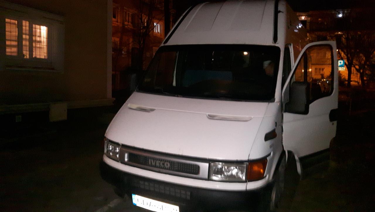 Biały samochód dostawczy Iveco z otwartymi drzwiami kierowcy, zaparkowany na zewnątrz budynku mieszkalnego w nocy, widoczny znak drogowy P.