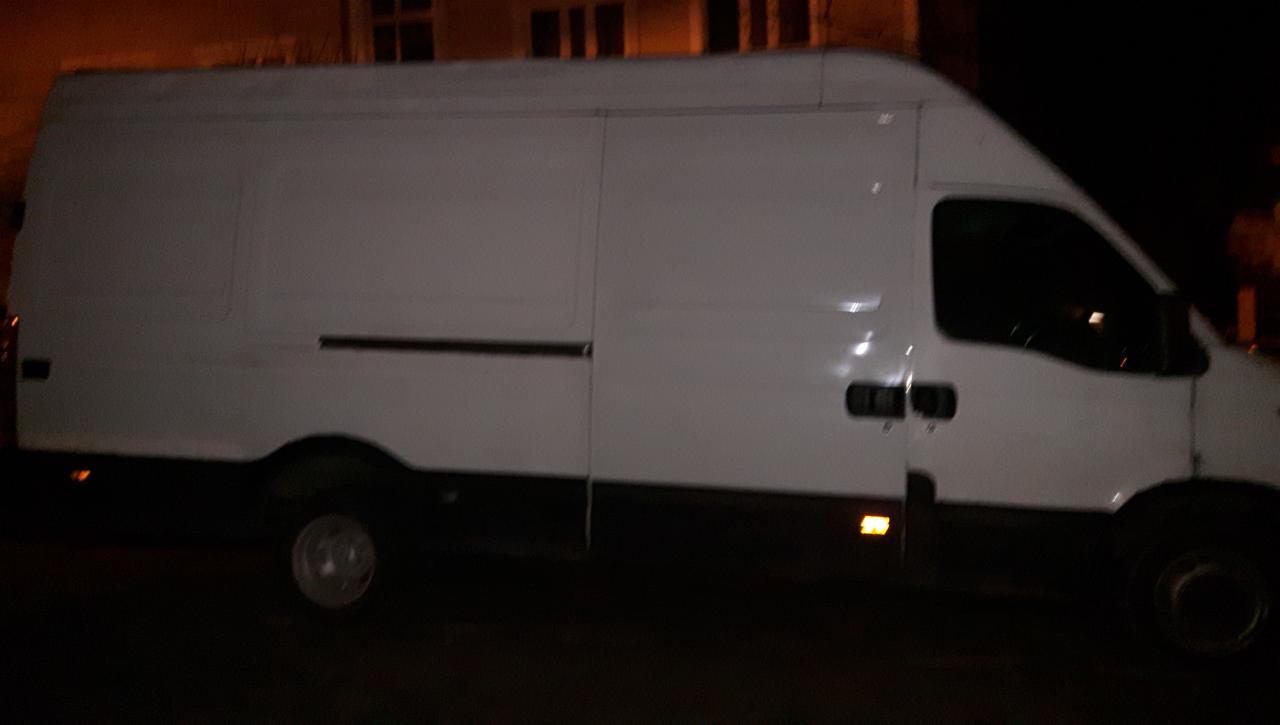 Biały samochód dostawczy zaparkowany nocą, widoczne światła pozycyjne i zarys budynku w tle.