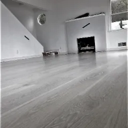 Wykończona podłoga drewniana w jasnoszarym odcieniu, widok na białe ściany z kominkiem i nietypowym okrągłym oknem, zabezpieczona taśmą malarską przed dalszymi pracami.