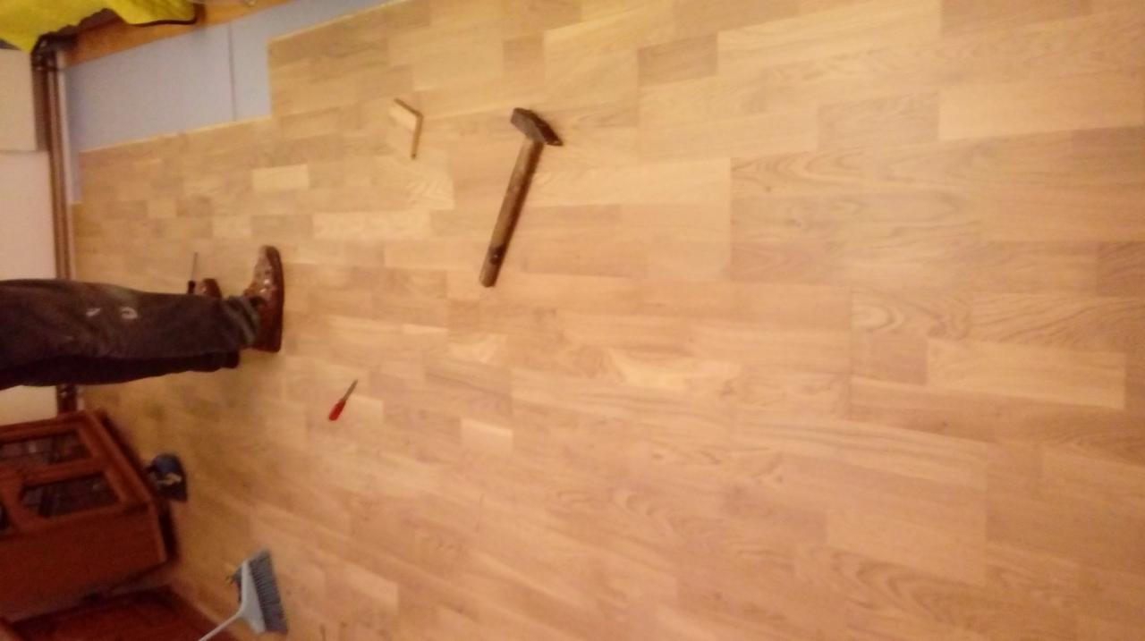 Fragment podłogi z parkietu w trakcie układania, widoczny młotek, fragment nogi w brązowym bucie i narzędzia.