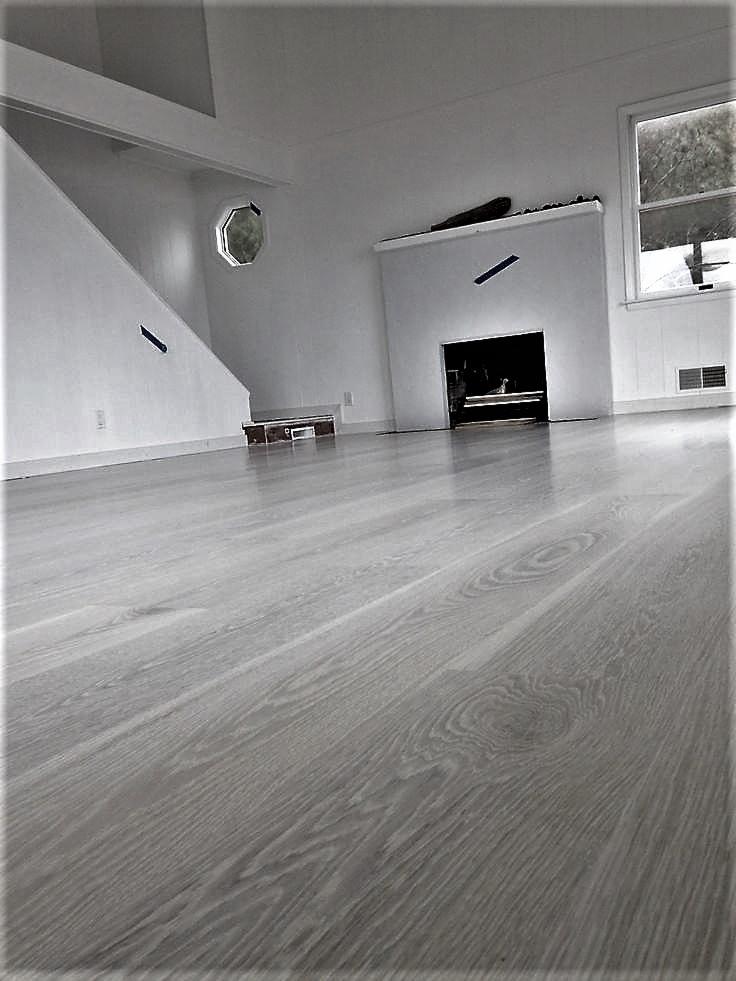 Wykończona podłoga drewniana w jasnoszarym odcieniu, widok na białe ściany z kominkiem i nietypowym okrągłym oknem, zabezpieczona taśmą malarską przed dalszymi pracami.