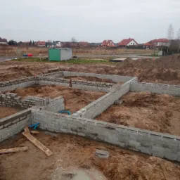 Wykopane fundamenty domu z częściowo wymurowanymi ścianami z bloczków betonowych, widoczne niebieskie rury instalacyjne, teren budowy w pochmurny dzień.