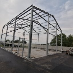 Robspaw - Stalowa konstrukcja hali w trakcie budowy, widoczne elementy szkieletu, betonowa podstawa, dwóch pracowników i częściowo widoczny samochód dostawczy.