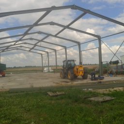 Robspaw - Konstrukcja stalowa hali w trakcie montażu na betonowej płycie, z widocznym traktorem JCB i pracownikiem przy spawaniu, na tle pola i pochmurnego nieba.