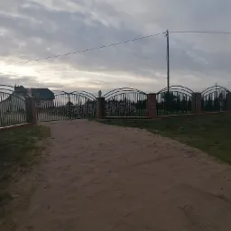 Kuta brama wjazdowa z ceglanymi słupkami i dekoracyjnymi elementami w kształcie lwa, prowadząca na posesję z widocznym stosem drewna i domem w tle, częściowo zasłonięta przez zaparkowany samochód.