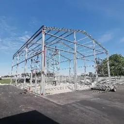 Stalowa konstrukcja hali w trakcie budowy, widoczne rusztowania i przygotowane płyty betonowe na podłożu, błękitne niebo w tle.