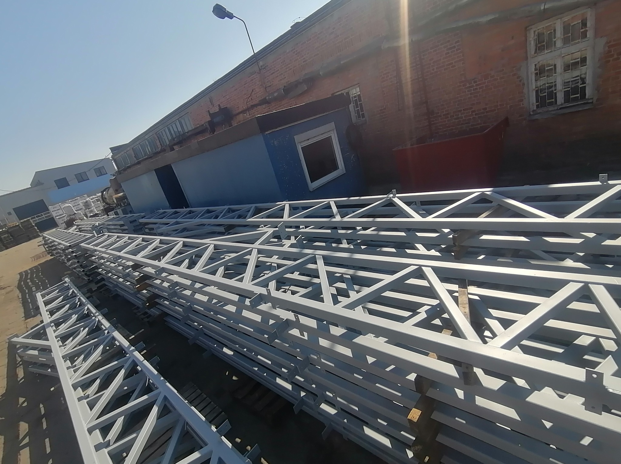 Stos konstrukcji stalowych z profili zamkniętych, pomalowanych na szaro, na tle ceglanego budynku. Elementy do budowy hali.