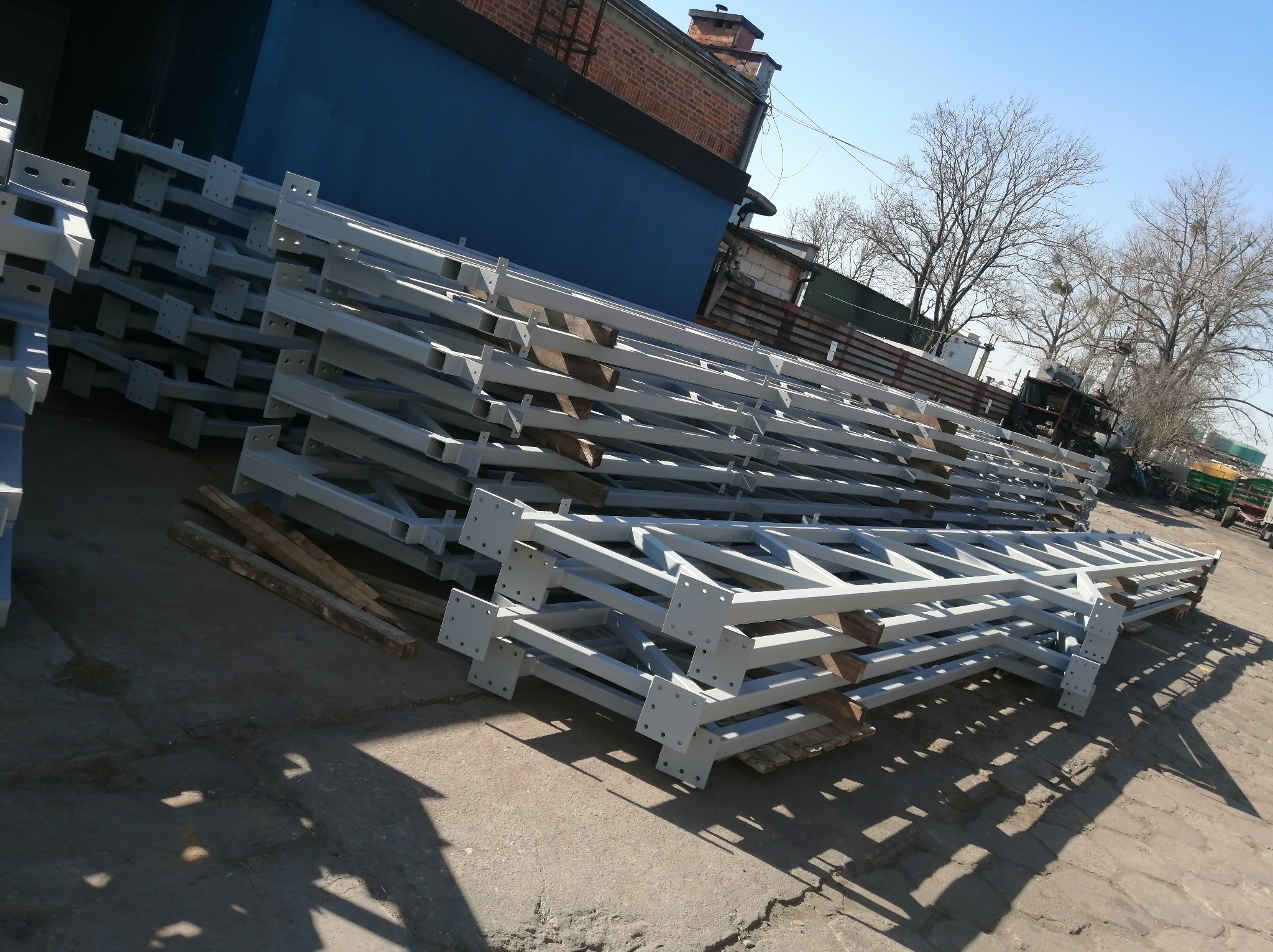 Stalowe elementy konstrukcyjne, pomalowane na szaro, ułożone w stos na placu budowy. Widoczne otwory montażowe i spawane połączenia. W tle budynek i drzewa.