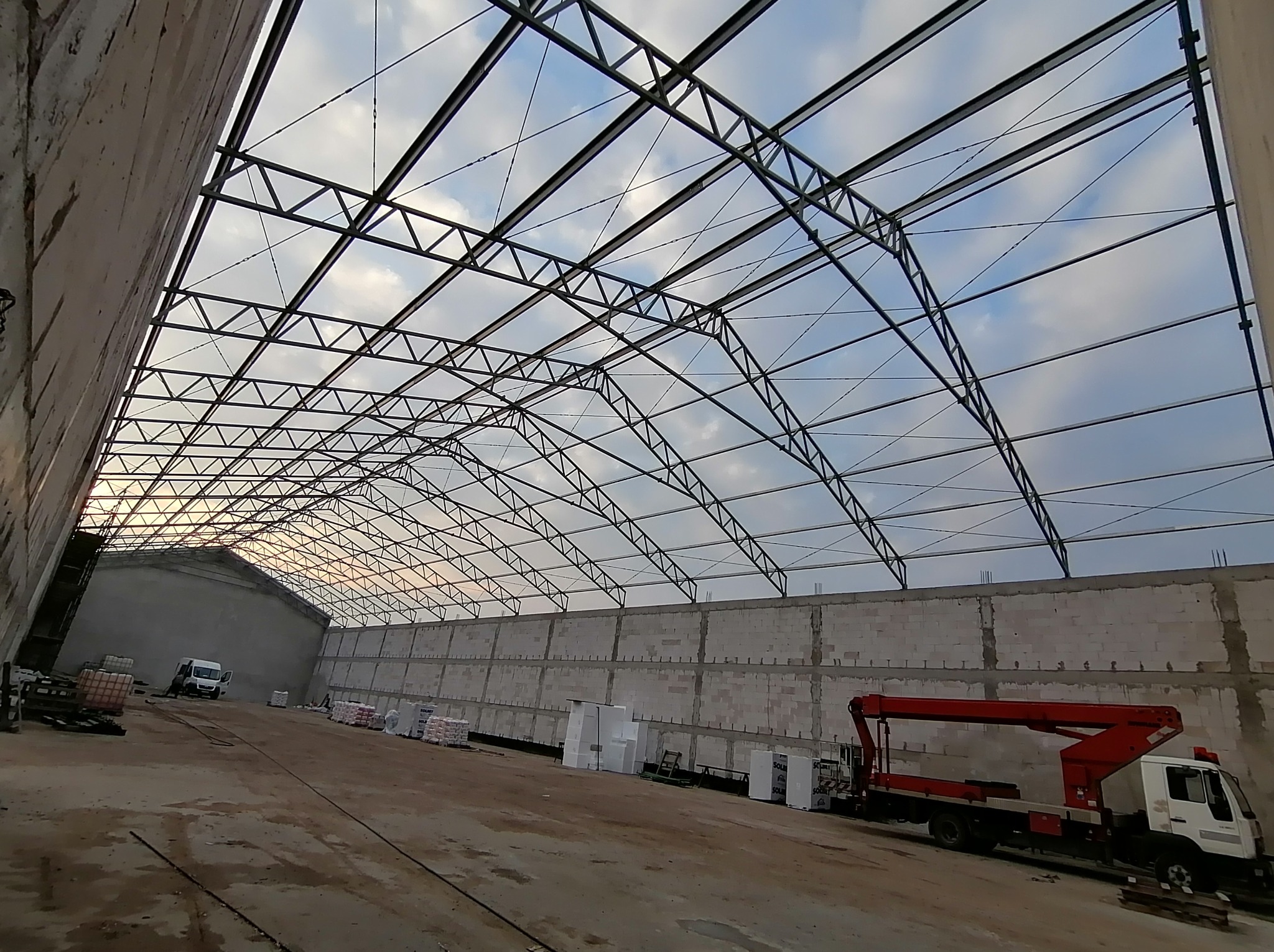 Wnętrze hali w trakcie budowy z widoczną konstrukcją stalową dachu i ścianami z betonu komórkowego. Na podłodze materiały budowlane i podnośnik koszowy.