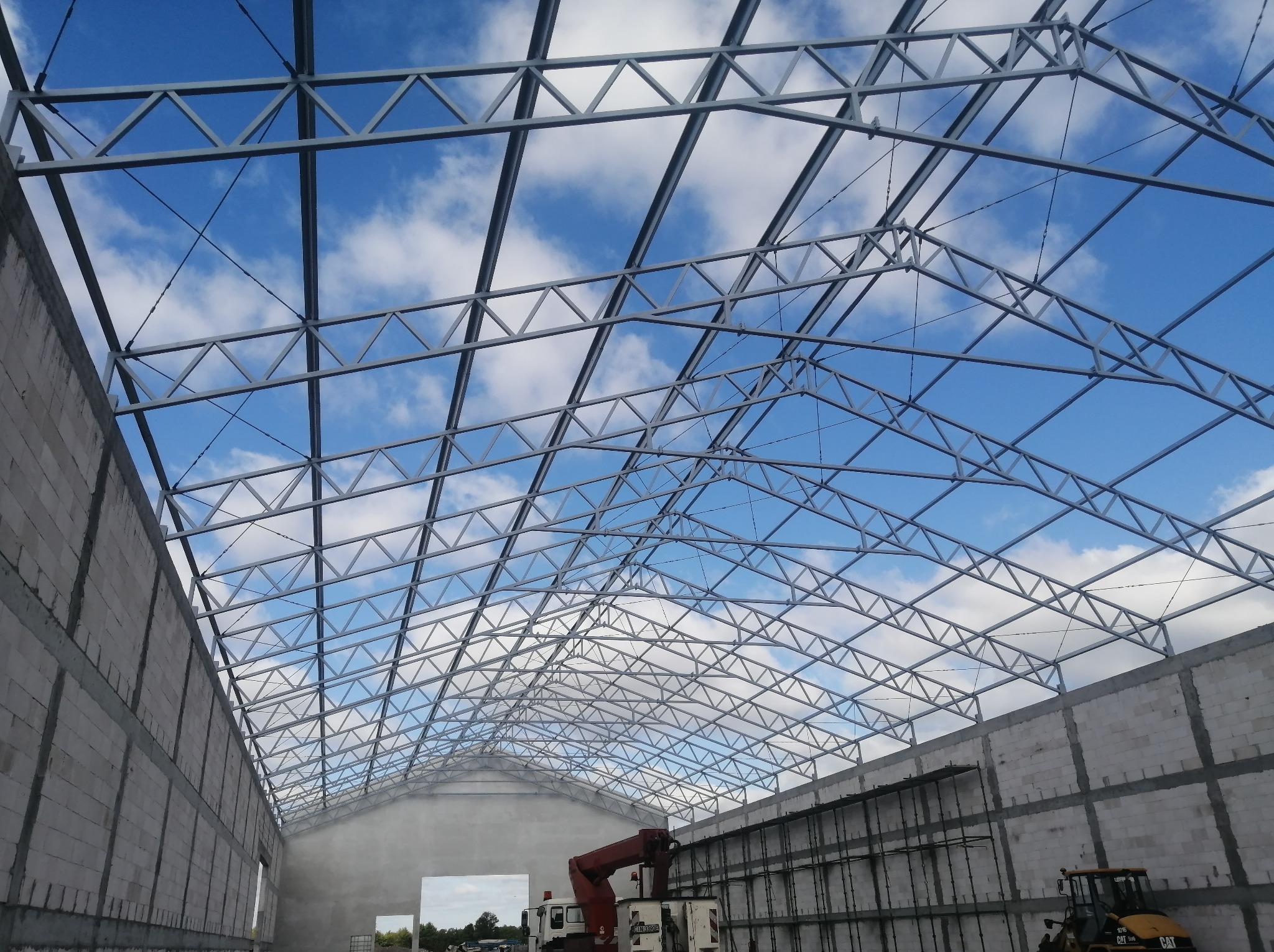 Wnętrze hali w budowie z widoczną stalową konstrukcją dachu na tle błękitnego nieba z chmurami. Ściany z betonu komórkowego, sprzęt budowlany w tle.