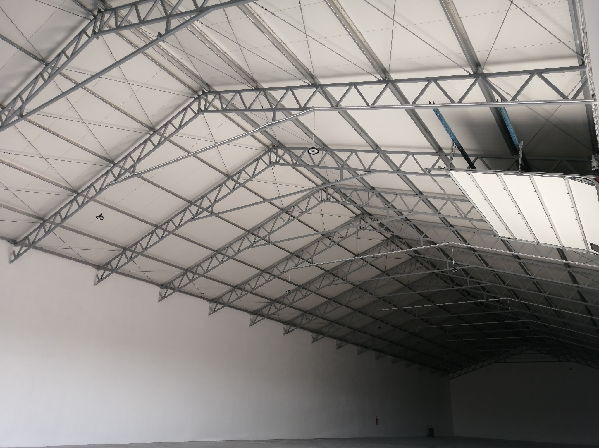 Wnętrze hali z widoczną konstrukcją stalową dachu, oświetleniem i białymi ścianami. Perspektywa od dołu ukazuje przestrzeń i geometrię konstrukcji.