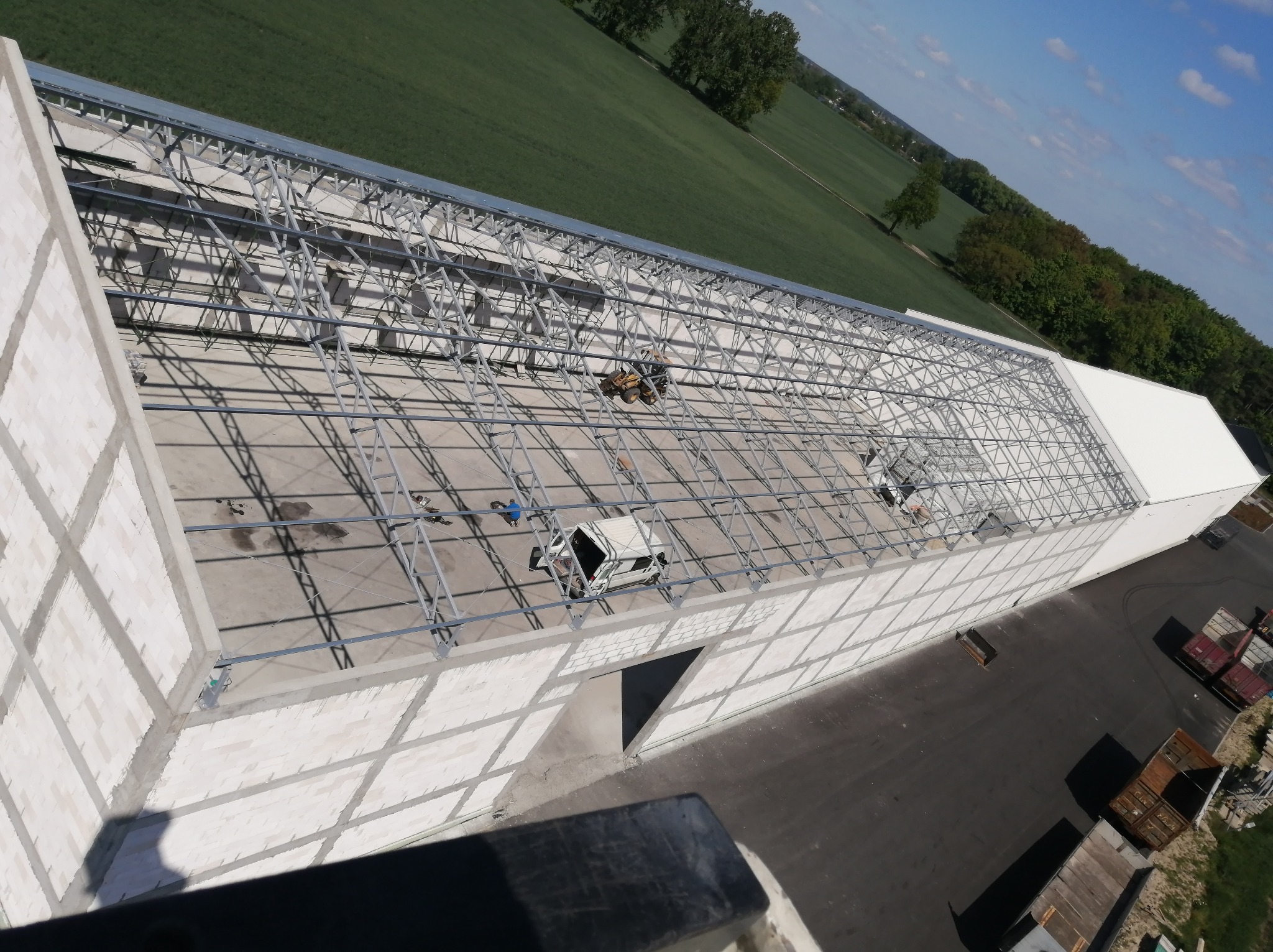 Budowa hali w toku: widok z góry na konstrukcję stalową z białymi ścianami, maszyny budowlane na dachu, zielone pola w tle. Nietypowy kąt ujęcia.