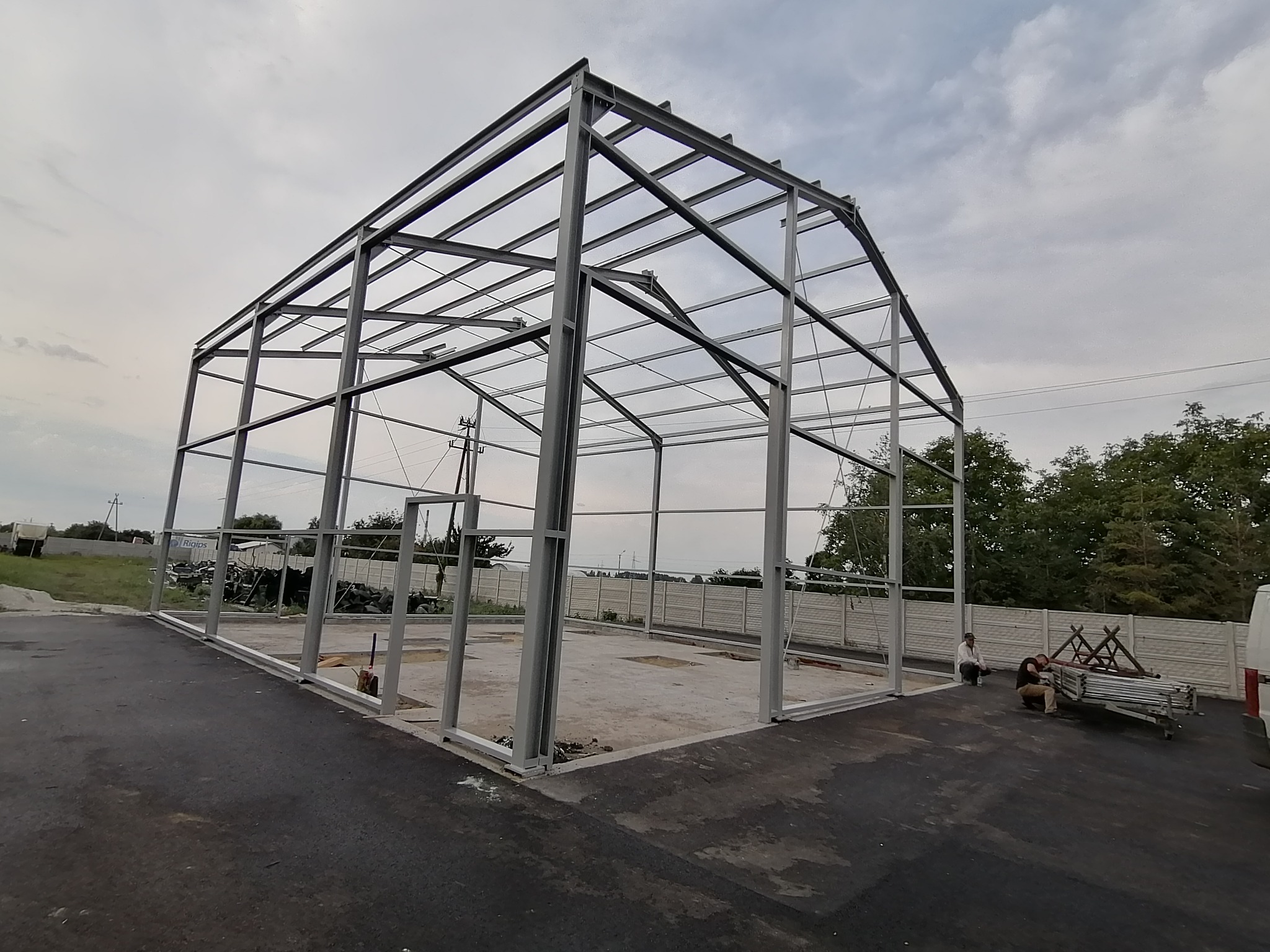 Stalowa konstrukcja hali w trakcie budowy, widoczne elementy szkieletu, betonowa podstawa, dwóch pracowników i częściowo widoczny samochód dostawczy.