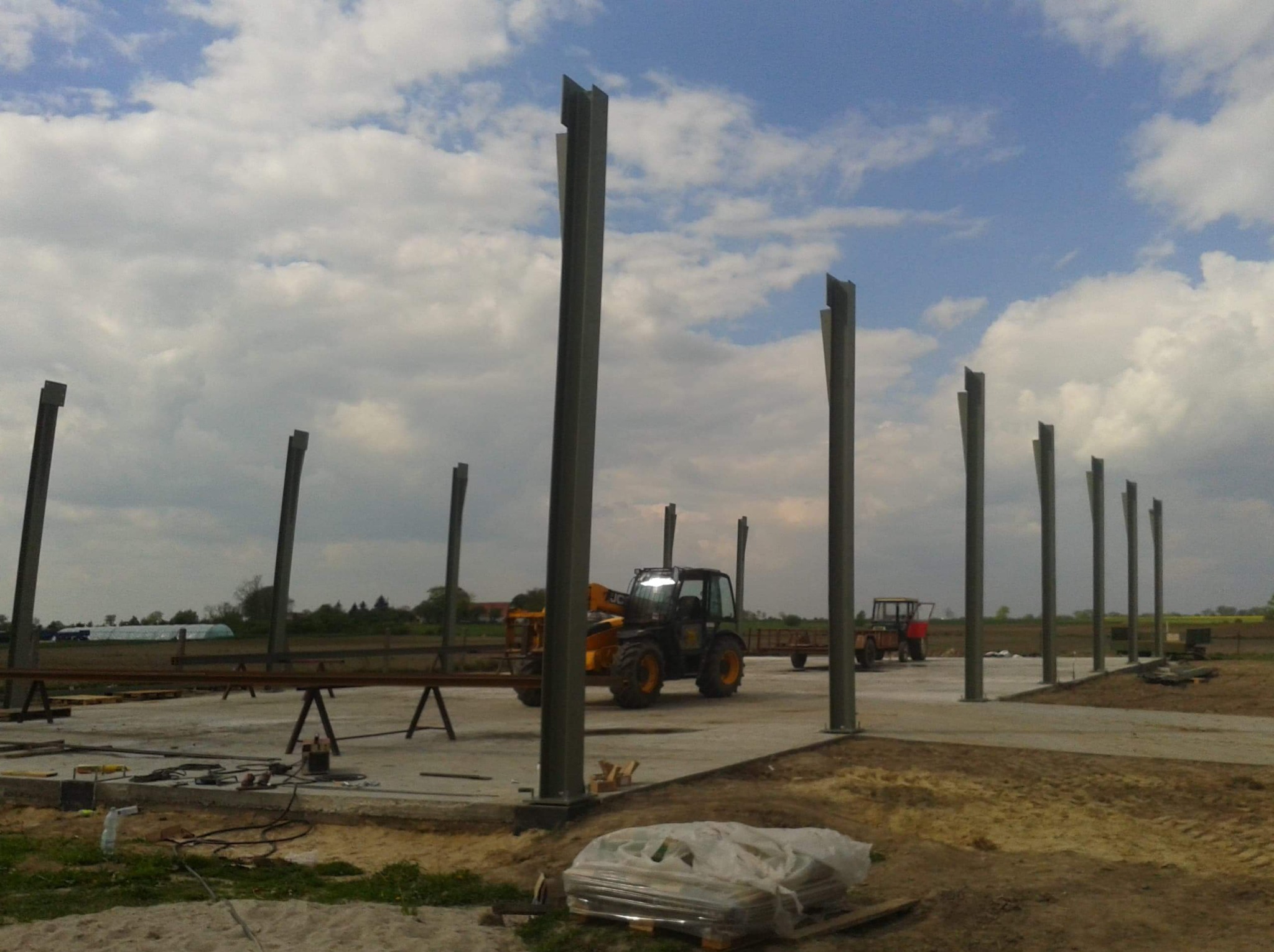 Montaż stalowych słupów hali na betonowej płycie fundamentowej, widoczny podnośnik teleskopowy i elementy konstrukcji. Prace w toku, pochmurne niebo.