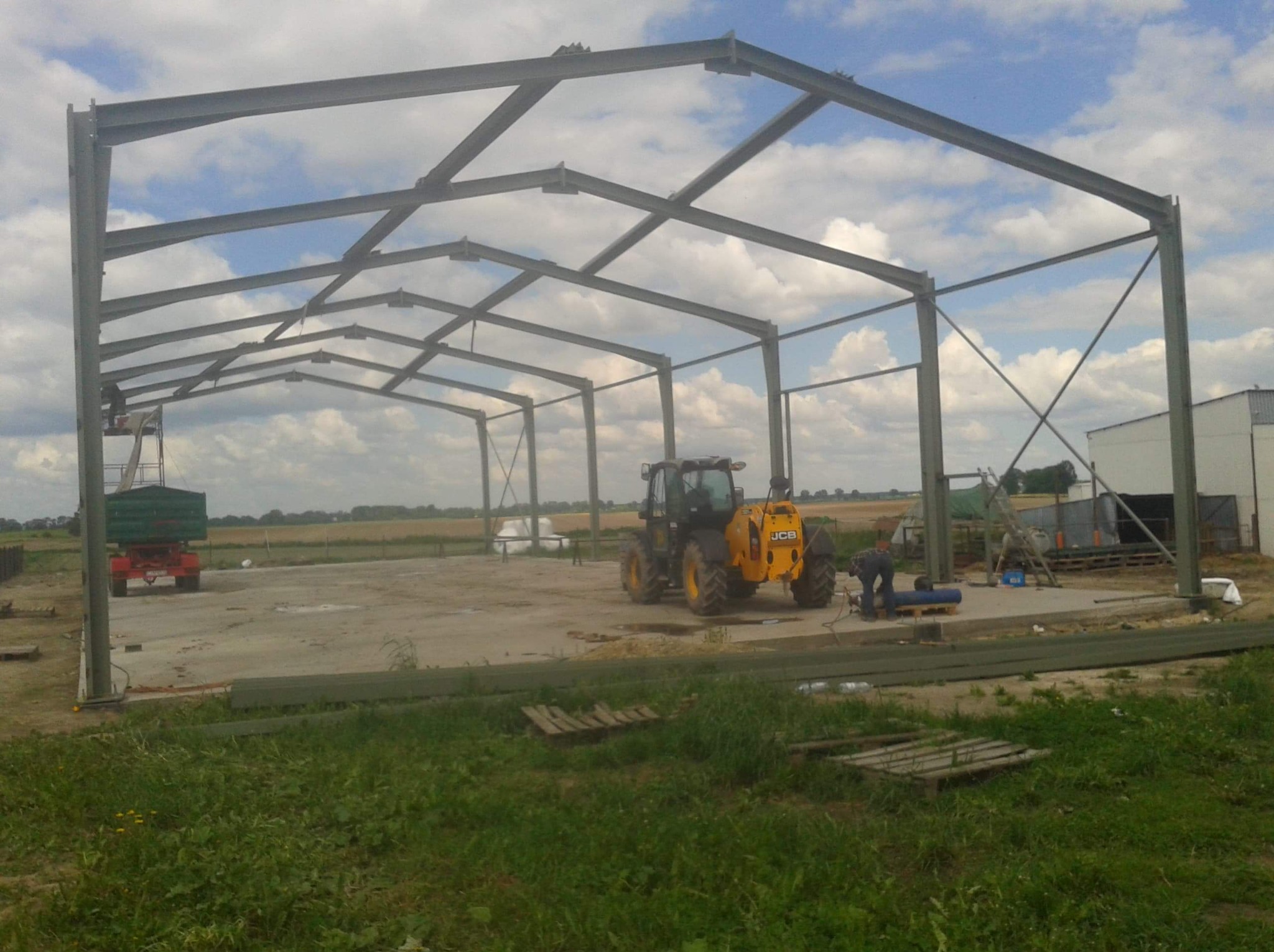 Konstrukcja stalowa hali w trakcie montażu na betonowej płycie, z widocznym traktorem JCB i pracownikiem przy spawaniu, na tle pola i pochmurnego nieba.