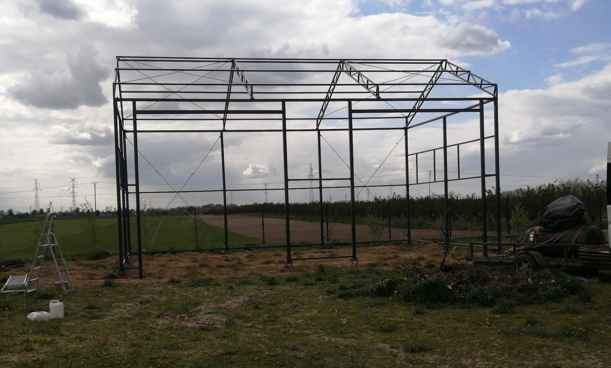 Stalowa konstrukcja szkieletowa hali w trakcie budowy, widoczna drabina i przyczepa w tle, pochmurne niebo.