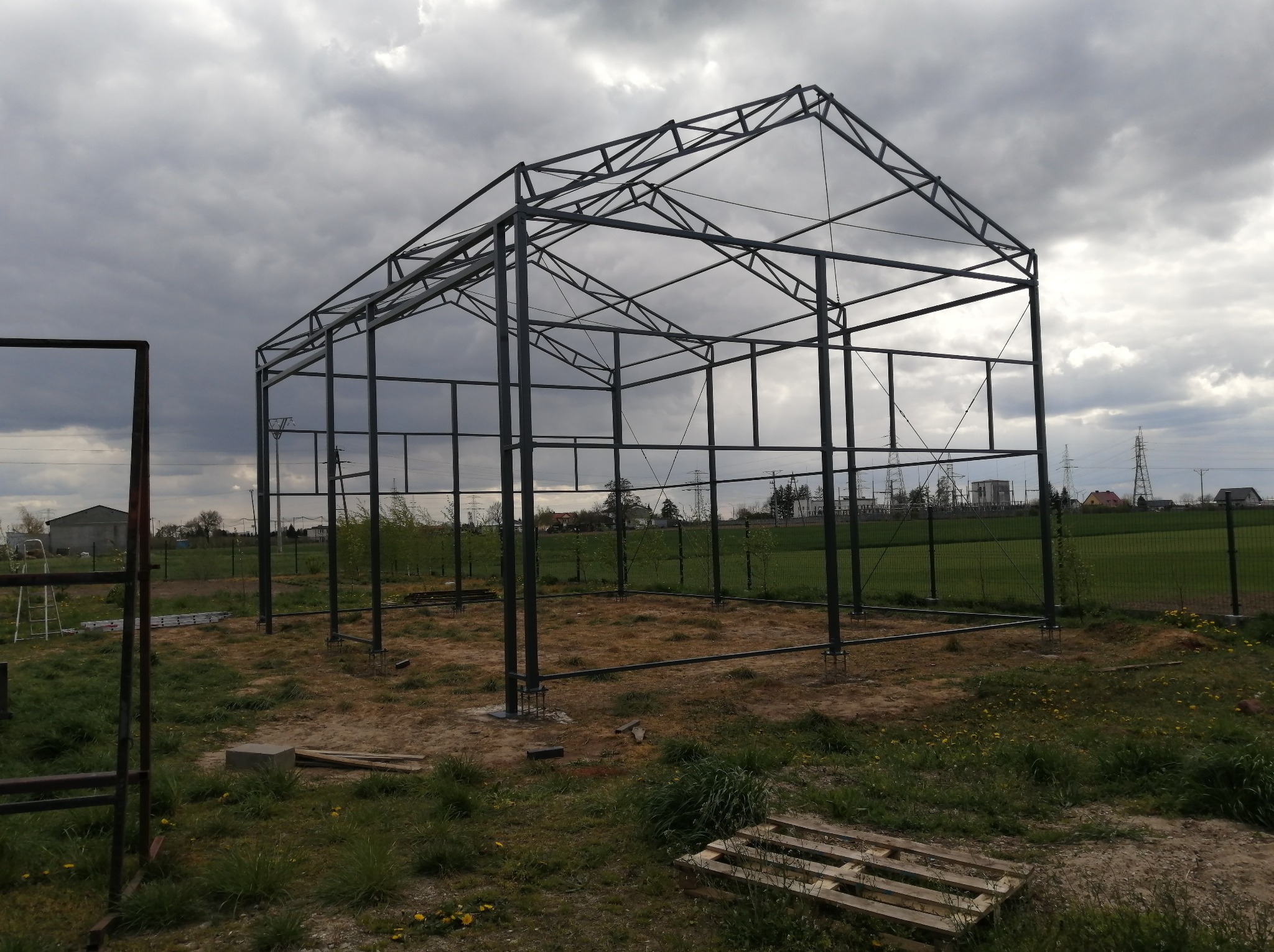 Konstrukcja stalowa hali w trakcie montażu, widoczne słupy i szkielet dachu na tle pochmurnego nieba. Paleta leży na ziemi.