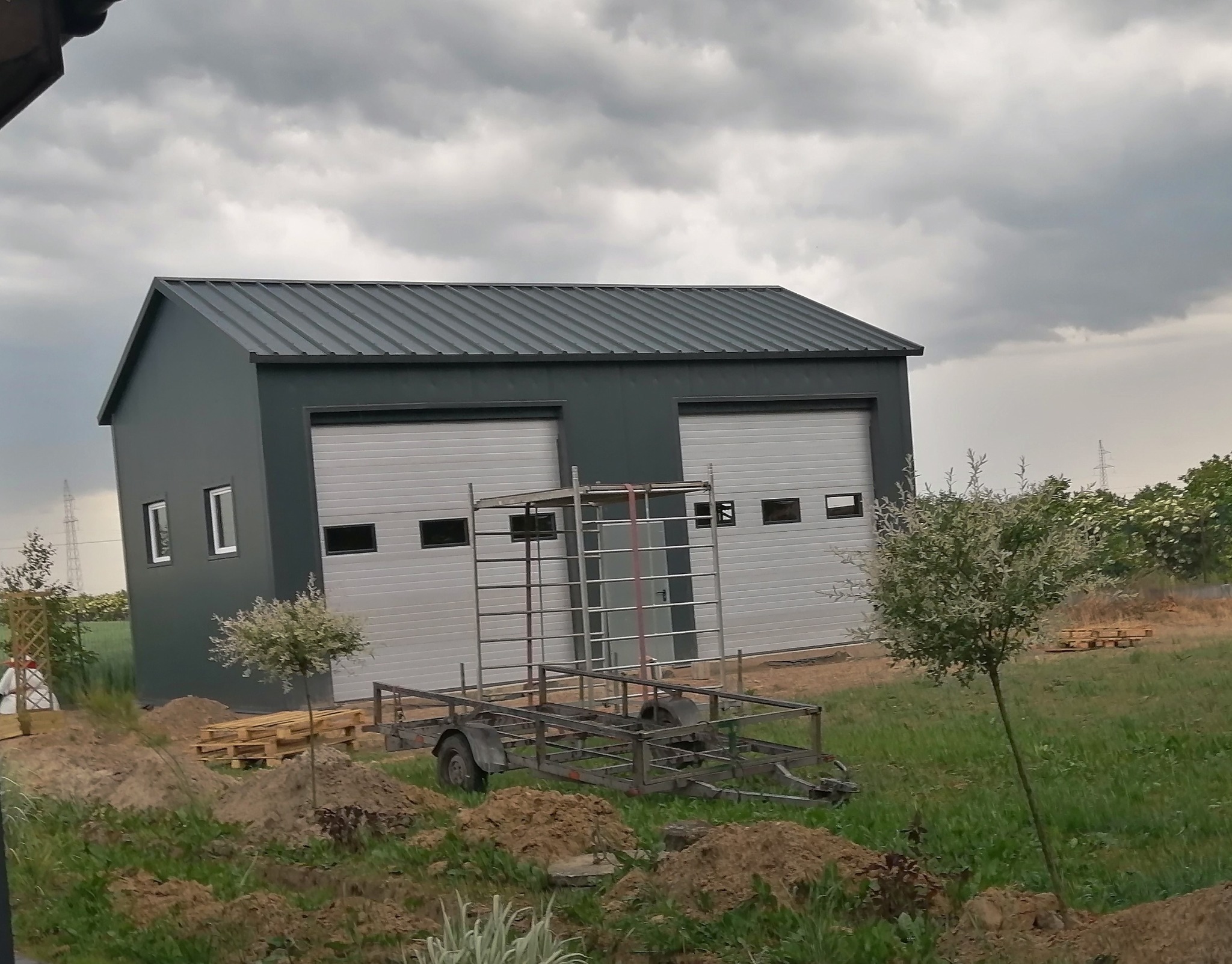 Nowoczesna hala z dwoma bramami garażowymi, widoczna z zewnątrz, z metalową konstrukcją i przyczepką na pierwszym planie, w tle pochmurne niebo.
