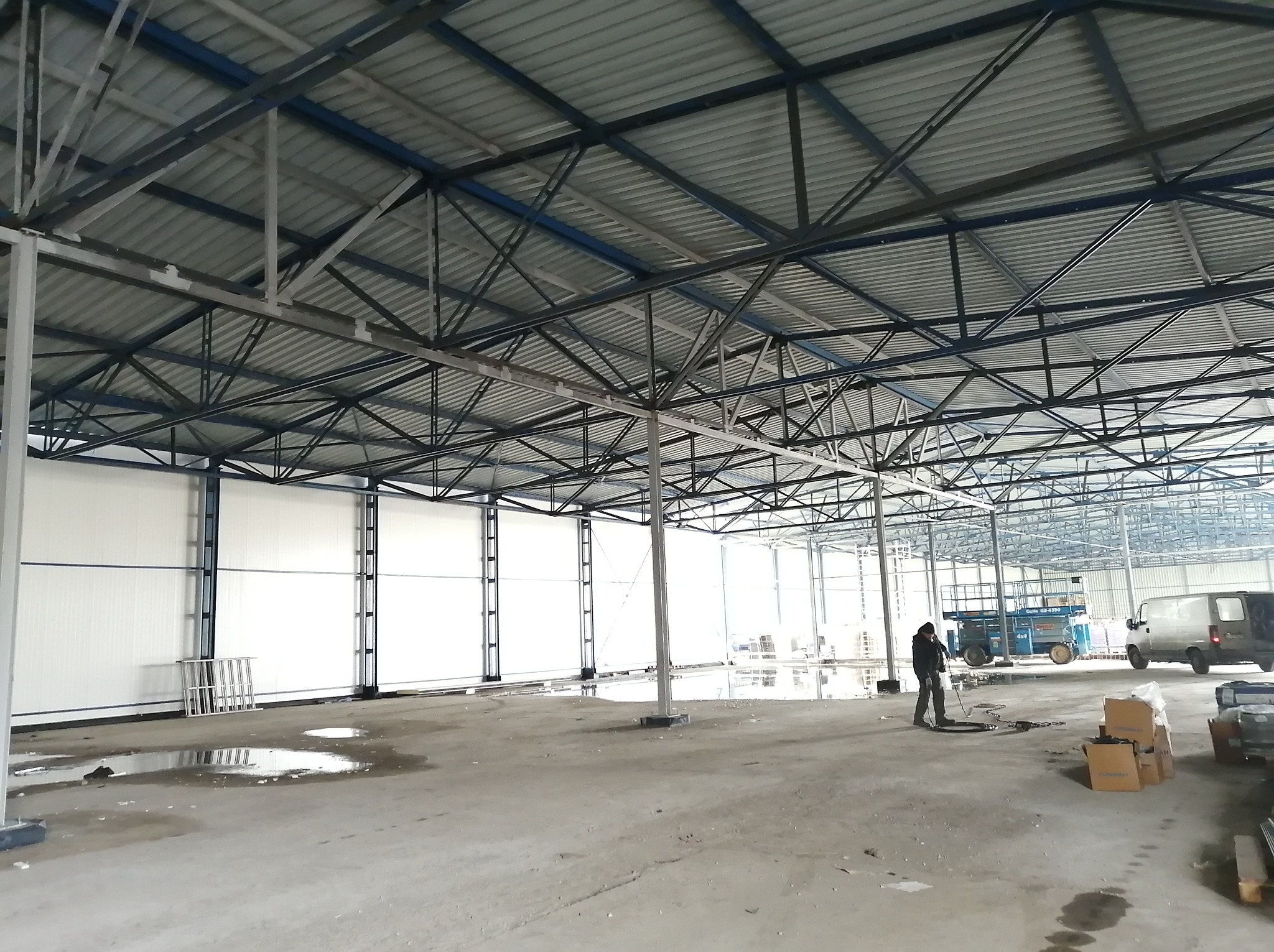 Wnętrze hali w trakcie budowy z widoczną konstrukcją stalową dachu, ścianami z paneli i pracownikiem. Na podłodze widoczne kałuże i materiały budowlane.