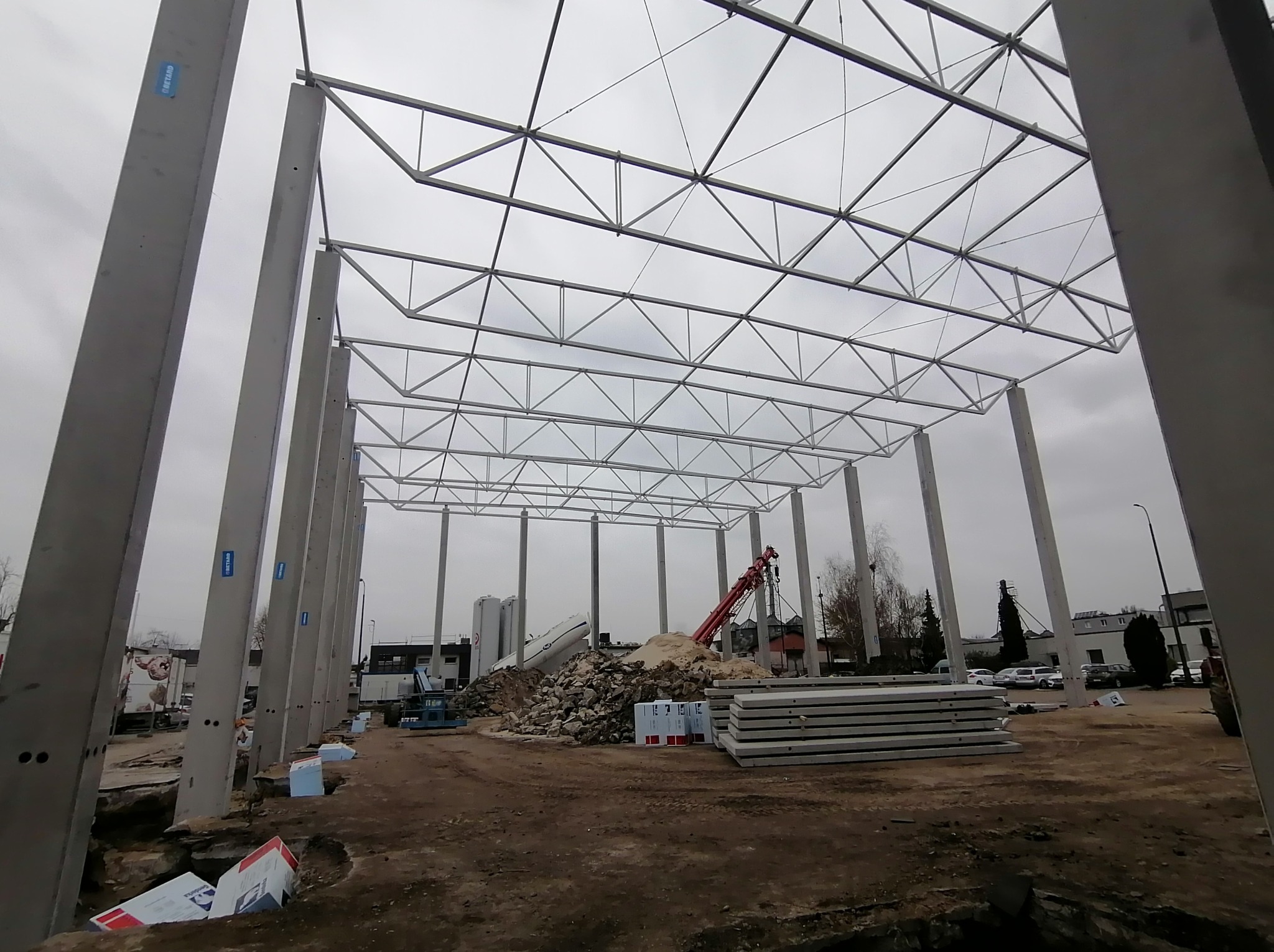 Wnętrze budowanej hali z widocznymi betonowymi słupami, stalową konstrukcją dachu i stertą gruzu. W tle dźwig i budynki. Pochmurne niebo.
