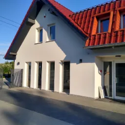 Jasny budynek z czerwoną dachówką, widoczne trzy wysokie okna, drzwi wejściowe i brama wjazdowa z samochodem w tle.