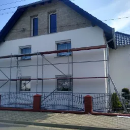 Dom jednorodzinny w trakcie malowania elewacji na biało, z widocznymi rusztowaniami i fragmentem ogrodzenia z metaloplastyki.