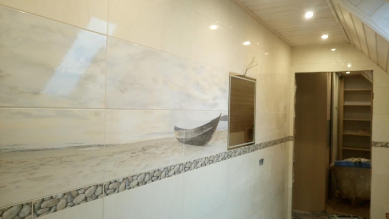 Wykończona łazienka z płytkami imitującymi pejzaż morski z łodzią na plaży, listwa dekoracyjna z motywem kamieni, biała podsufitka z oświetleniem punktowym, widoczne wejście do garderoby z półkami.