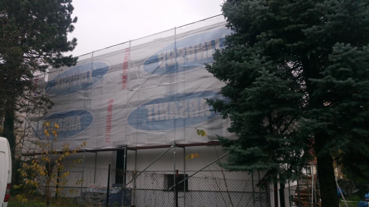 Budynek w trakcie prac elewacyjnych, osłonięty siatką z nadrukiem reklamowym na rusztowaniu, widoczne drzewa i fragment ogrodzenia.