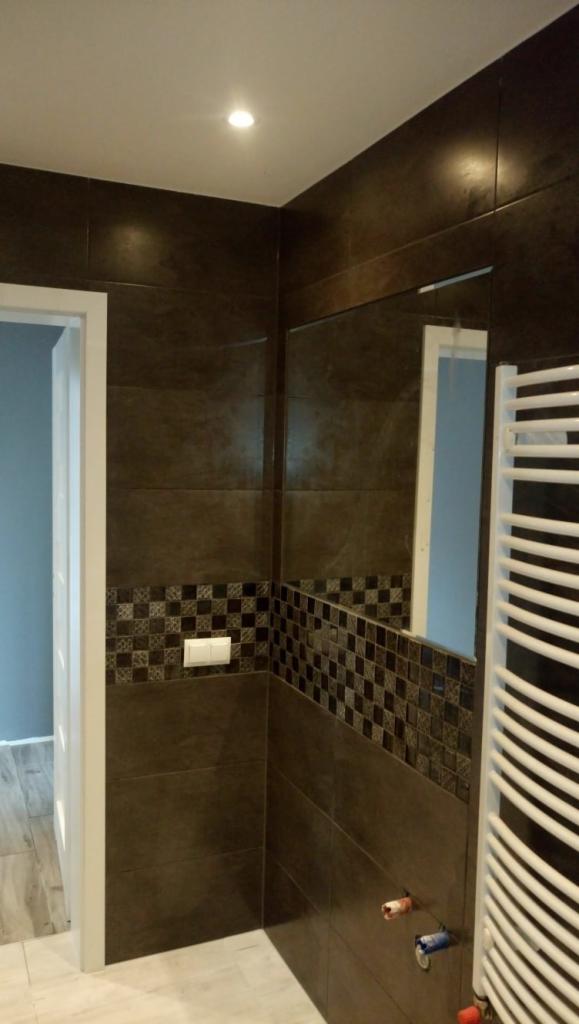 Ciemnobrązowe płytki w łazience z dekoracyjnym pasem mozaiki, biały grzejnik drabinkowy, włącznik światła i fragment lustra odbijający niebieską ścianę.