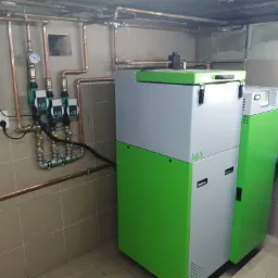 Nowoczesny piec CO z zielonymi akcentami, zainstalowany w pomieszczeniu z widoczną instalacją miedzianą, trzema pompami obiegowymi i manometrem.