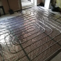 Rozłożona instalacja ogrzewania podłogowego na folii z siatką przed zalaniem w surowym wnętrzu. Widoczne rury grzewcze przymocowane do folii, ściany w trakcie remontu.