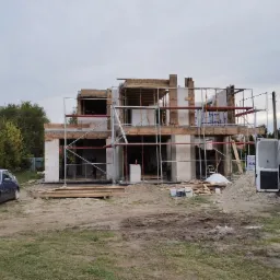 Surowy stan dwukondygnacyjnego domu z białego bloczka, widoczne rusztowania, drewniane szalunki, samochód osobowy i dostawczy na placu budowy.