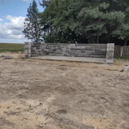 Wznoszone ściany parteru domu z bloczków betonowych na wylanej płycie fundamentowej, widoczne drzewa w tle i niebo z chmurami.