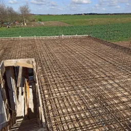 Zbrojenie płyty fundamentowej domu jednorodzinnego na tle zielonych pól i błękitnego nieba z chmurami.