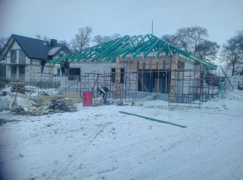 Wznoszenie ścian domu z białego bloczka w zimowej scenerii, widoczne rusztowania, drewniana konstrukcja dachu i pracujący robotnik w odblaskowej kamizelce.