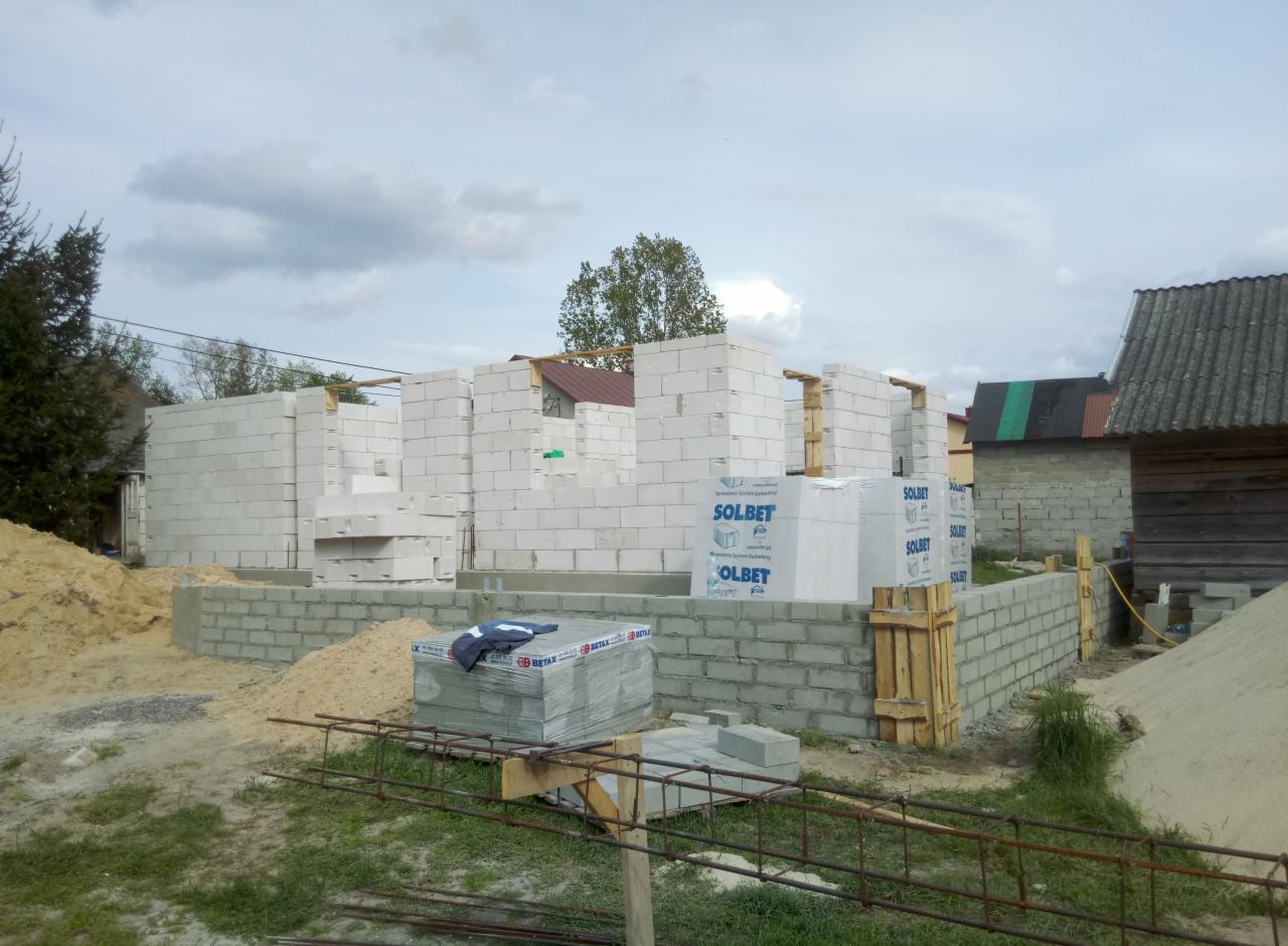 Wznoszenie ścian parterowego domu z bloczków Solbet na fundamencie z bloczków betonowych, widoczne palety z materiałem budowlanym i drewniane szalunki.