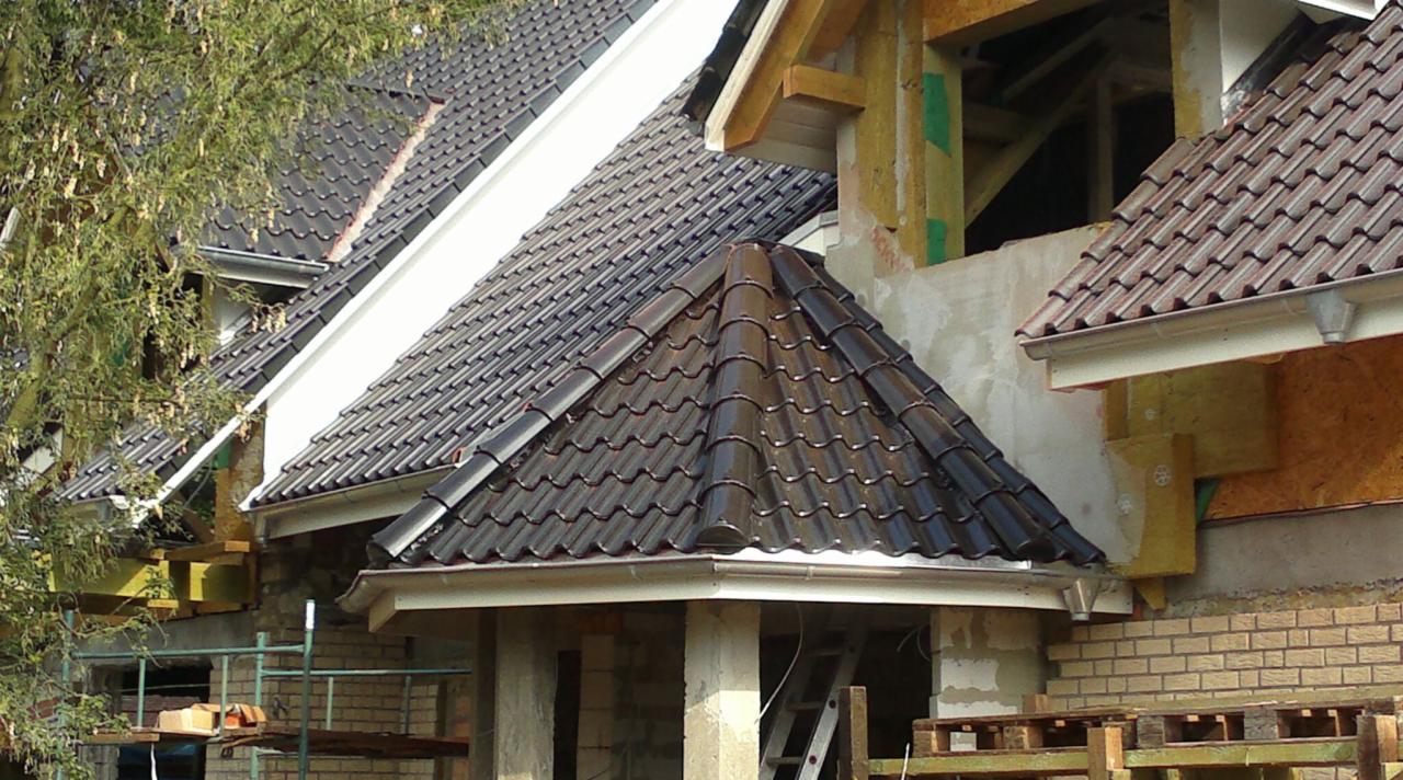 Nowe, błyszczące dachówki ceramiczne na wystającym fragmencie dachu domu w trakcie budowy. Widoczne rusztowania i materiały budowlane.