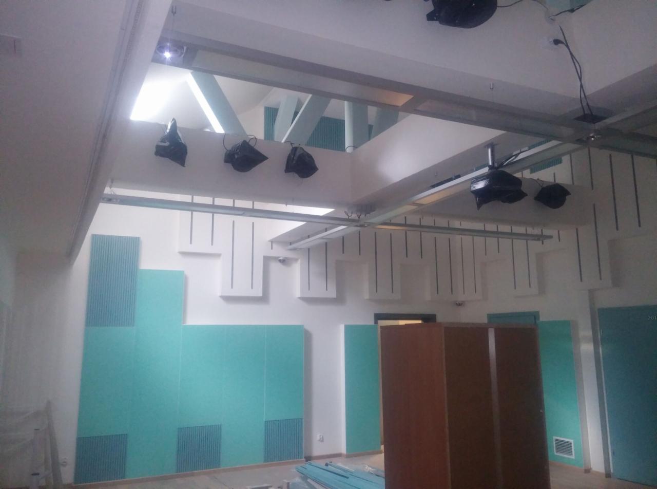 Wnętrze z elementami konstrukcji dachu: metalowe profile, drewniane belki, niebieskie panele akustyczne na ścianach. Widoczne oświetlenie i instalacje.